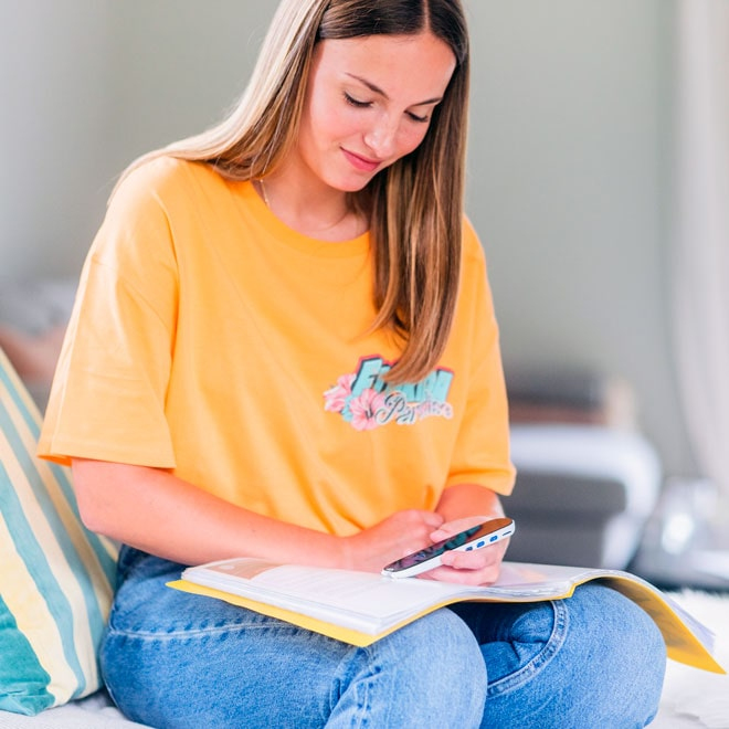 Eine junge Frau im orangefarbenen T-Shirt und Jeans benutzt einen Stift auf einem Handy über einem Buch.