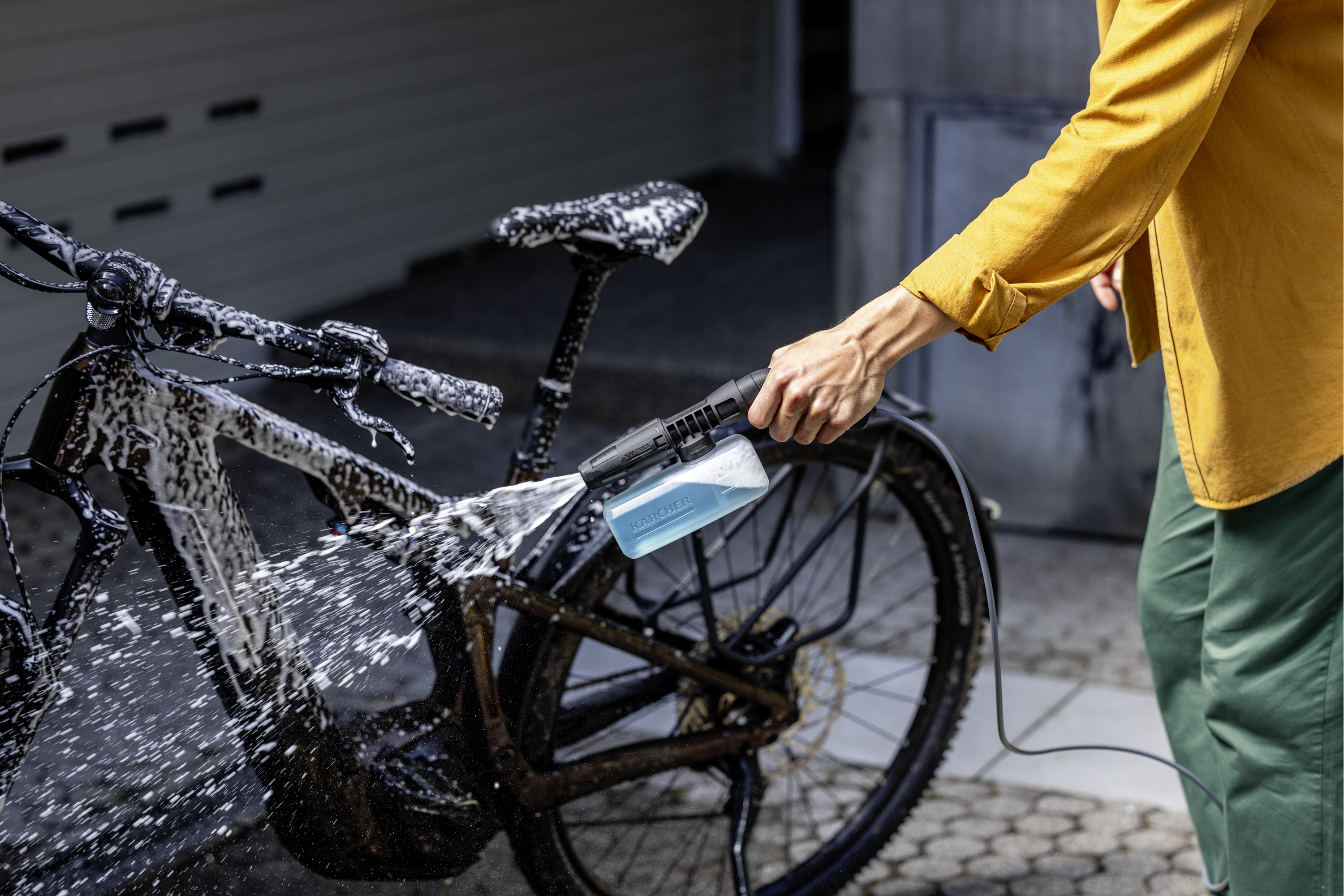 Persona con camicia gialla lava una bicicletta insaponata con idropulitrice e detergente blu.