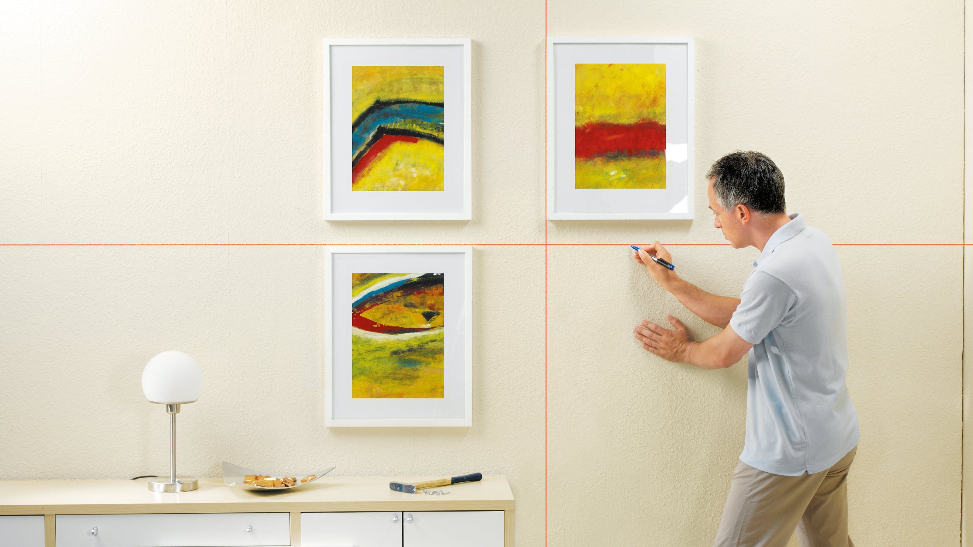 Mann markiert Wand mit Laser, Bilder hängen links. Tisch mit Lampe rechts.