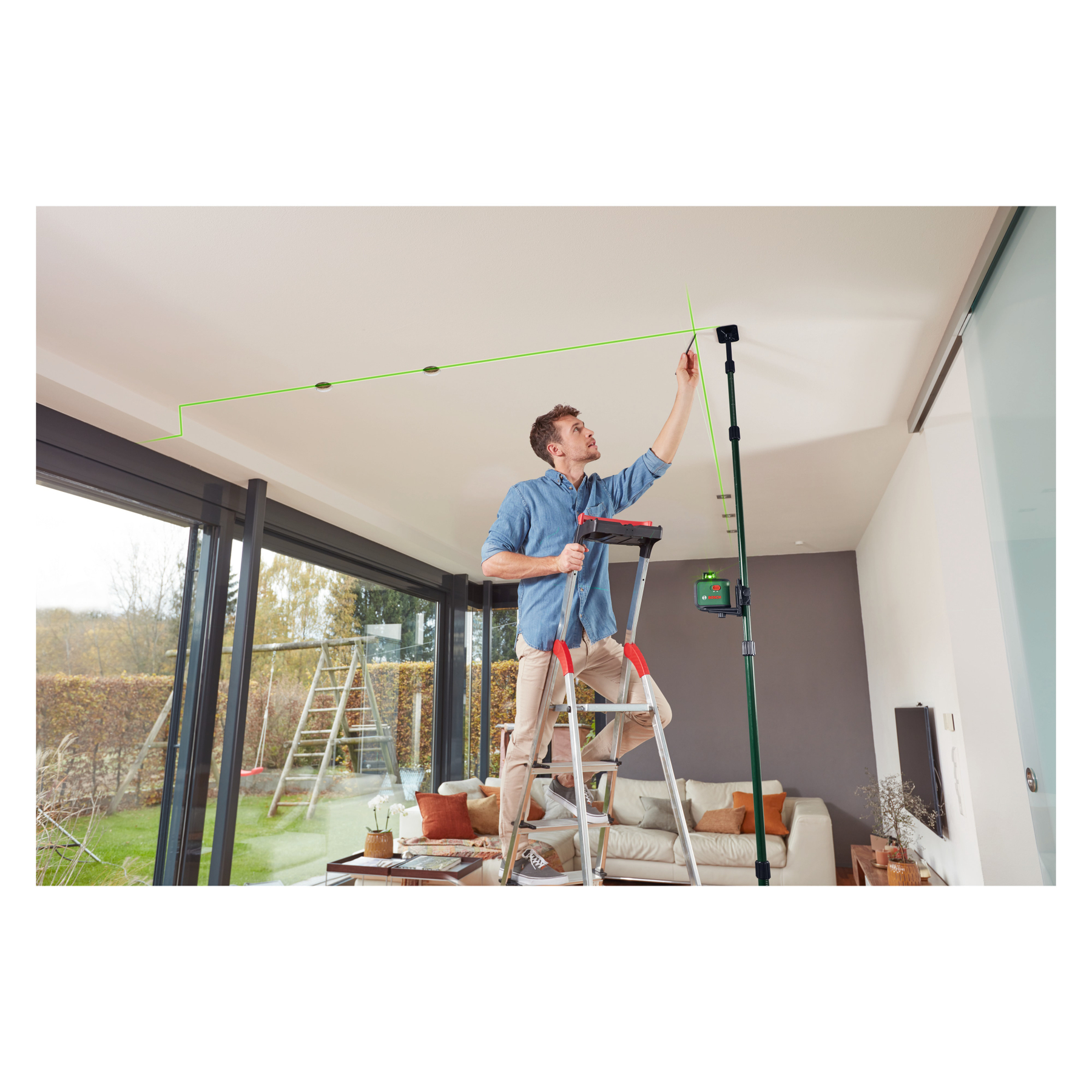 Mann auf Leiter richtet Laser an Decke aus. Wohnzimmer mit Fenstern.