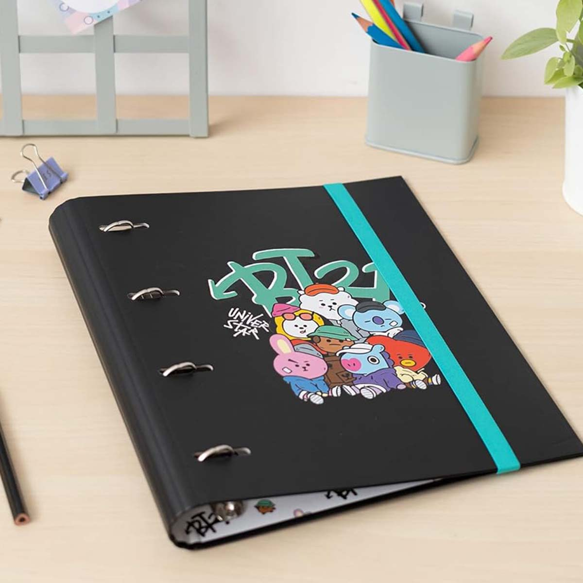 Schwarzer Ordner mit BT21-Figuren und türkisfarbenem Gummiband auf einem Schreibtisch.