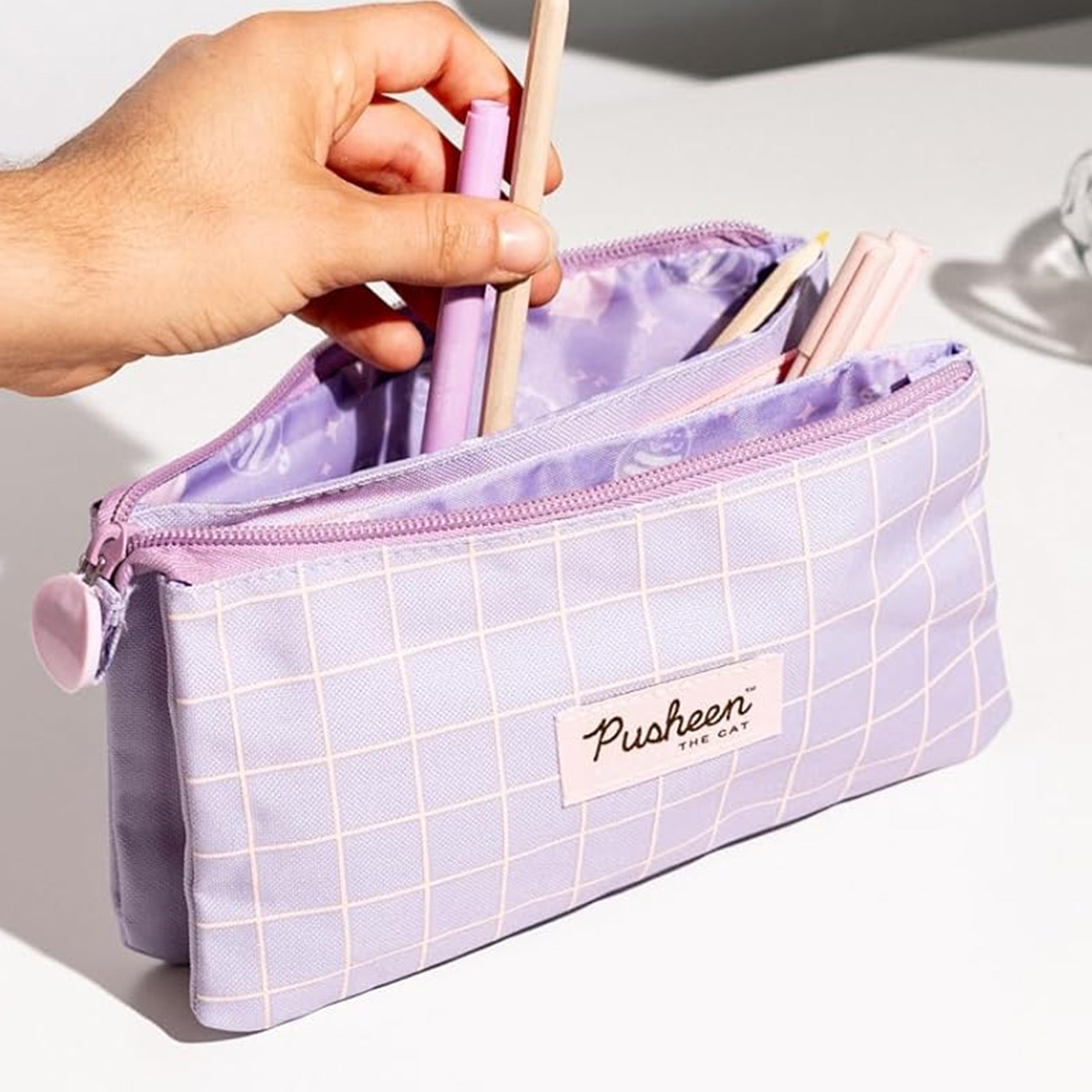 Eine Hand legt einen lila Stift in eine lavendelfarbene Federtasche mit kariertem Muster.