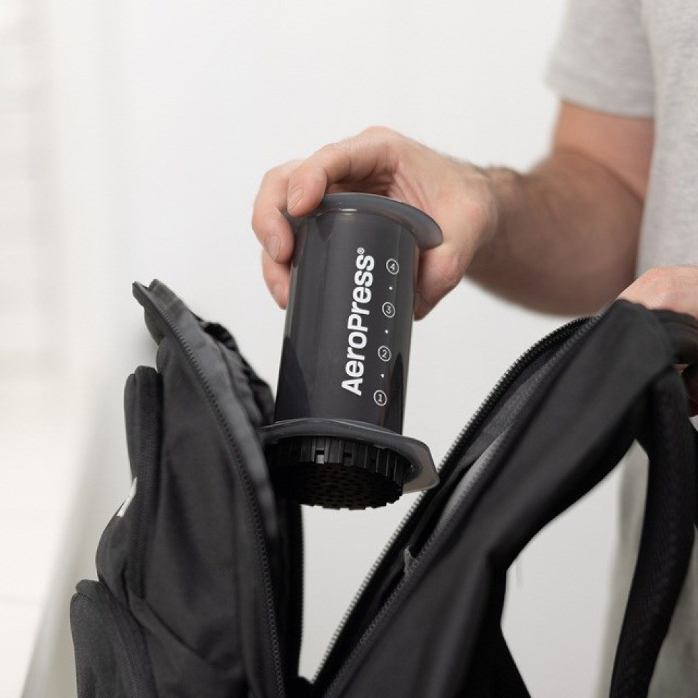 Una mano inserisce una caffettiera AeroPress grigia in uno zaino nero aperto.