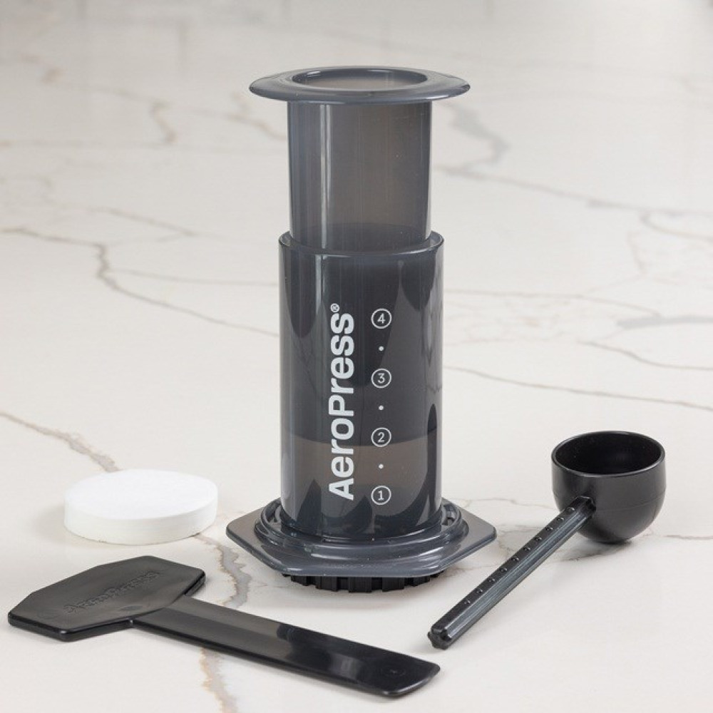 Macchina da caffè AeroPress con misurino, filtro e paletta su superficie in marmo.