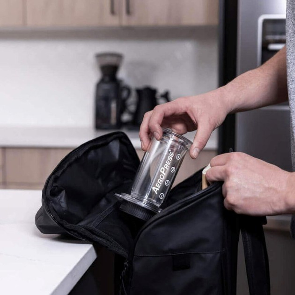 Ręka wkłada przezroczysty ekspres do kawy AeroPress do otwartego czarnego plecaka.