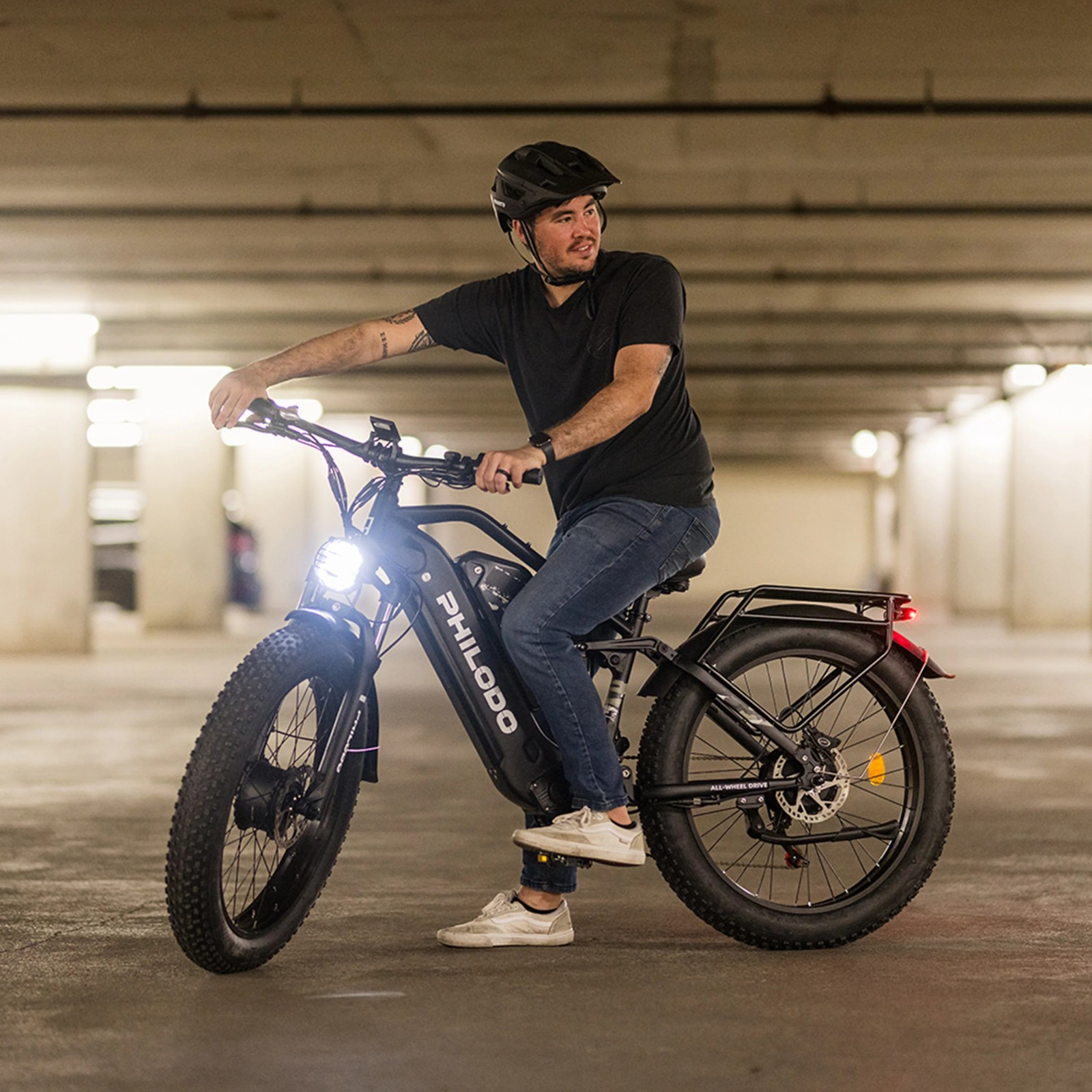 Mężczyzna w kasku jedzie na czarnym rowerze elektrycznym z szerokimi oponami w garażu.