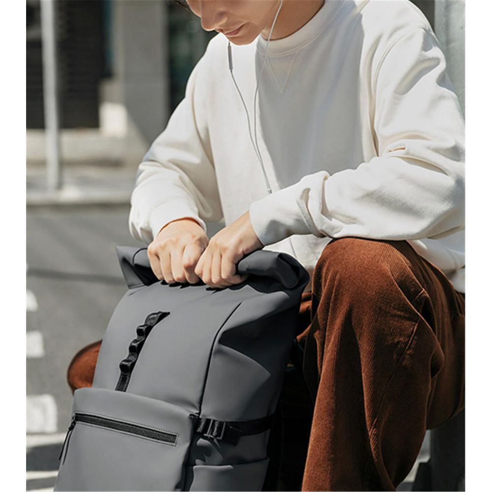 Hombre con suéter blanco y pantalones marrones con mochila gris, usa audífonos blancos.