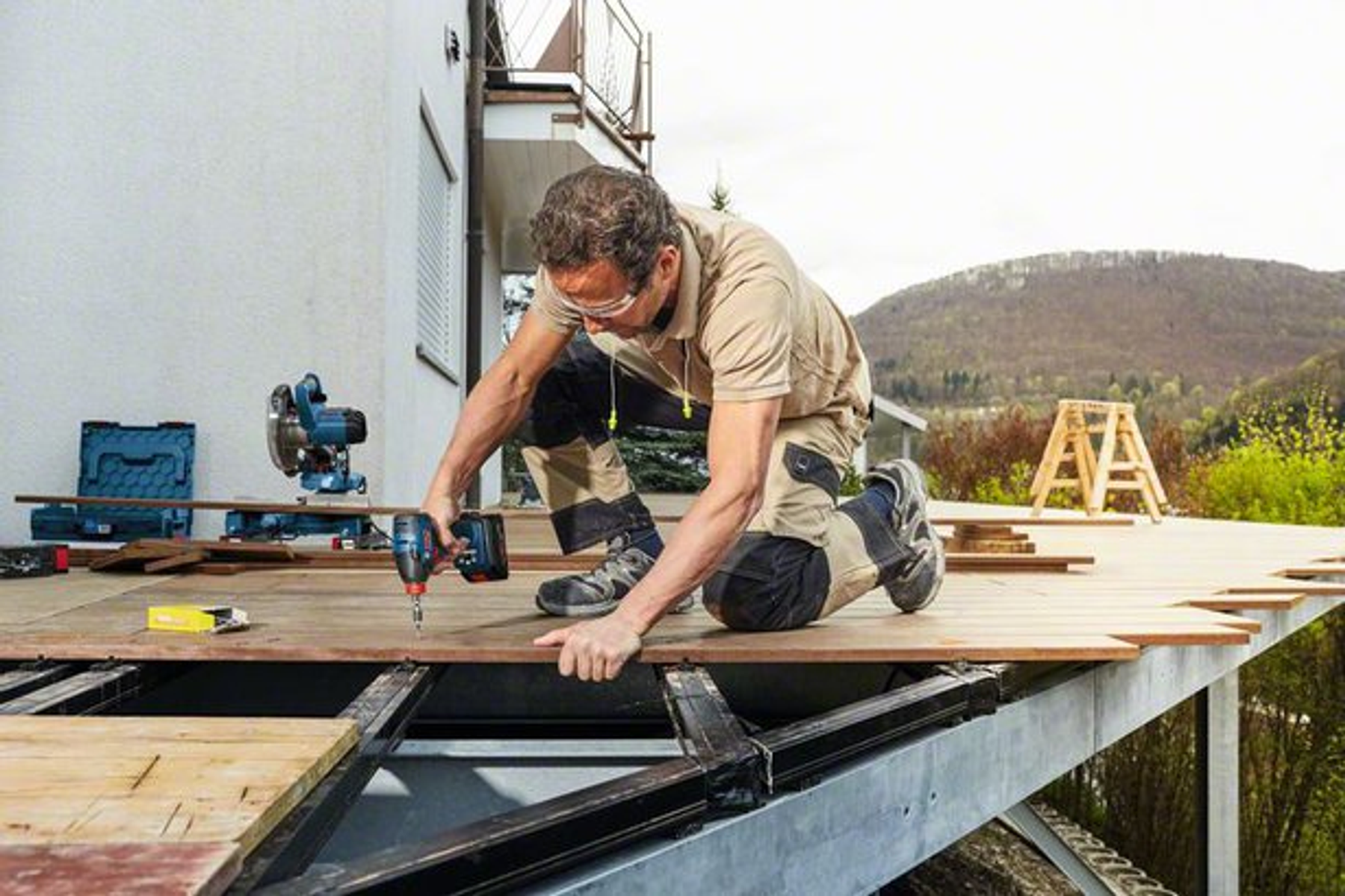 Ein Mann kniet auf einer Holzterrasse und schraubt mit einem Akkuschrauber.