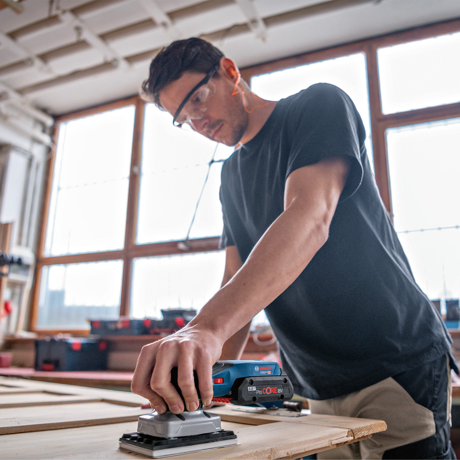 Mann mit Schutzbrille schleift Holz mit blauem Bosch-Schleifer in Werkstatt.