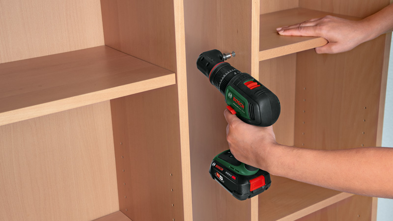Eine Hand benutzt einen Bohrer, um ein Regal in einem Holzschrank zu befestigen.