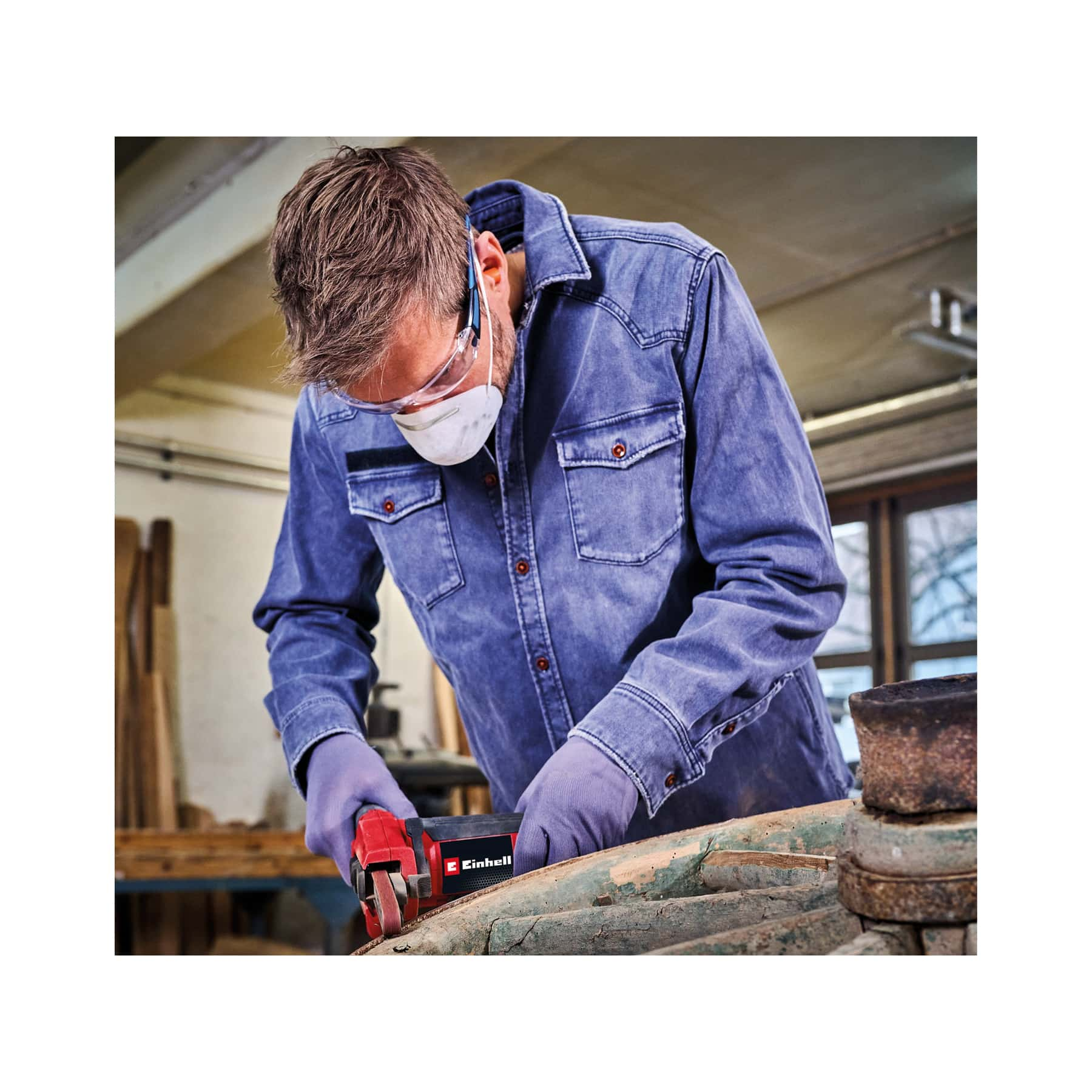 Mann mit Schutzbrille, Maske und Handschuhen benutzt Schleifgerät an Holz.