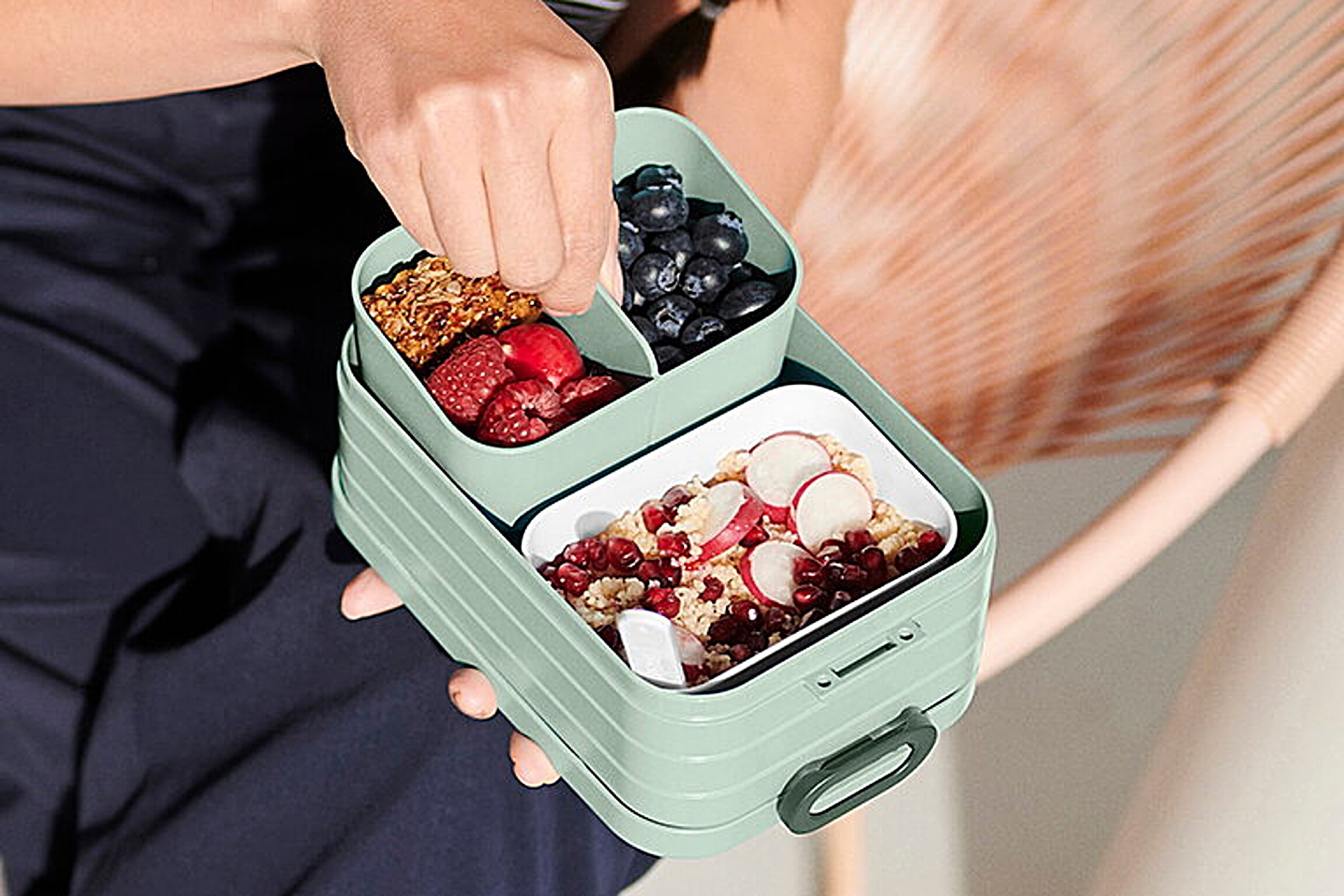 Eine Person öffnet eine grüne Brotdose mit Beeren, Müsli und Obst.