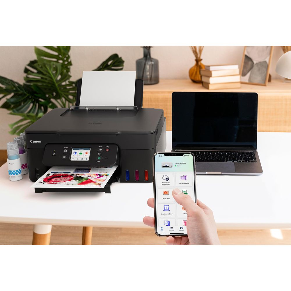 Ein Canon Drucker, Laptop und Handy mit Canon App stehen auf einem Schreibtisch. Fotos werden gedruckt.