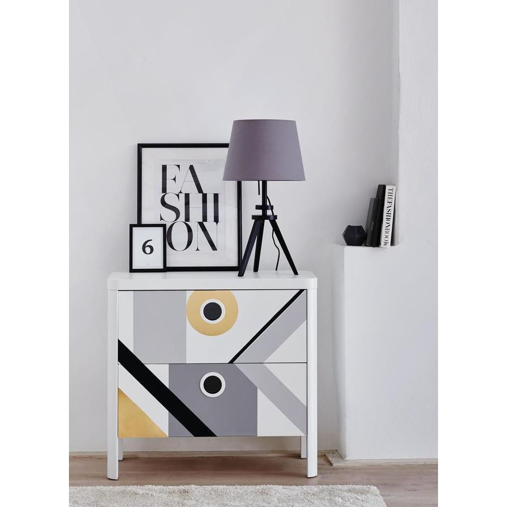 Weiße Kommode mit geometrischem Muster, Lampe, Buch und gerahmtem Bild.