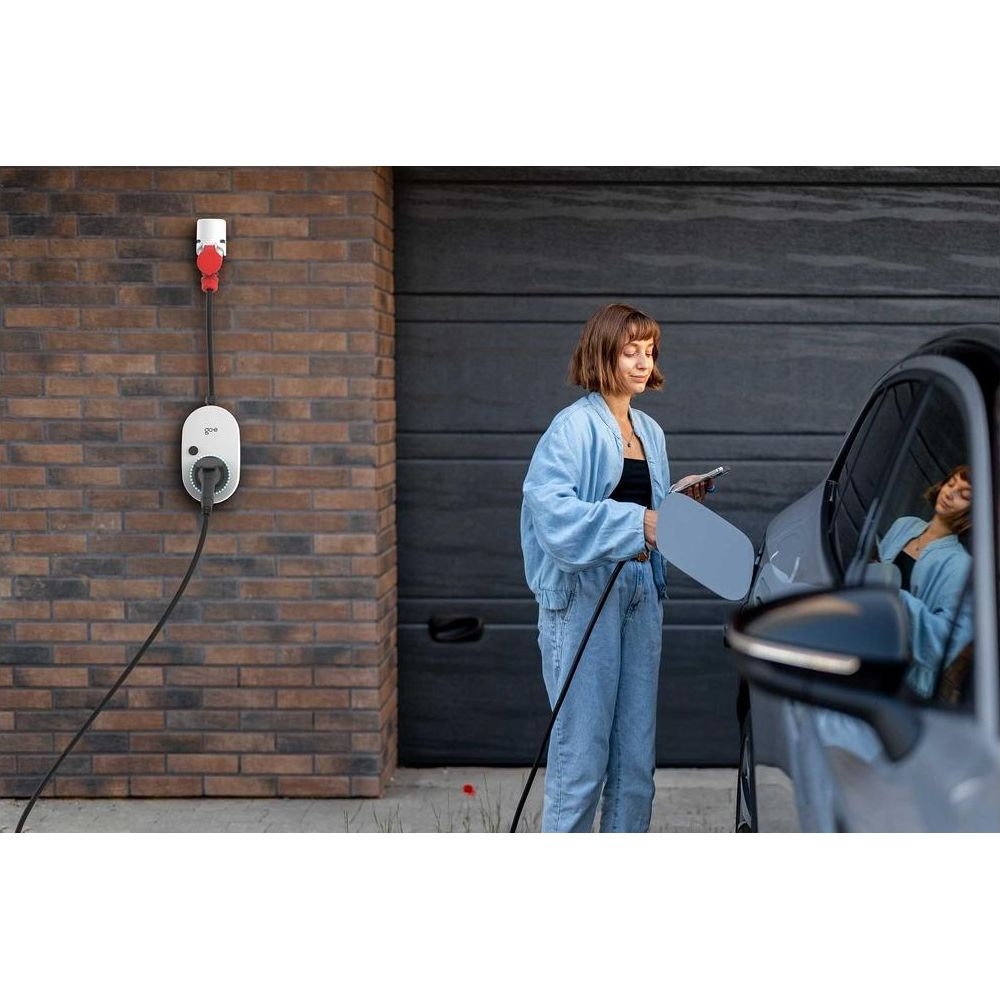 Frau lädt Elektroauto an Ziegelwand-Ladestation, hält Handy.