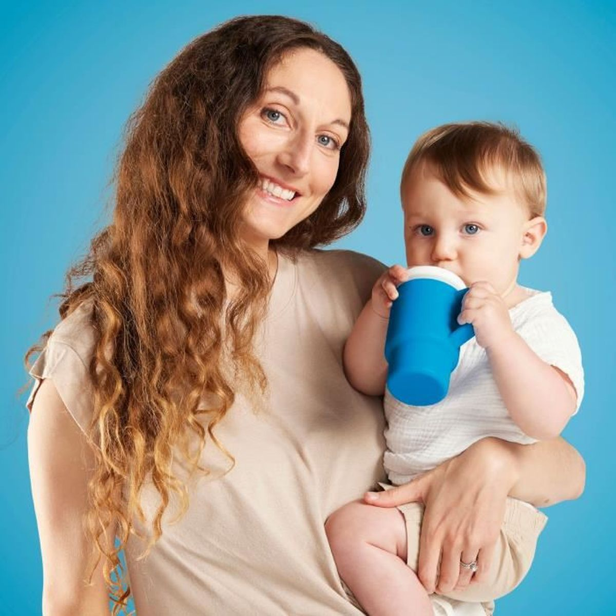 Eine Frau hält ein Baby, das aus einem blauen Becher trinkt. Blauer Hintergrund.