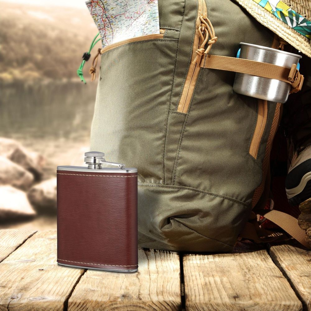 Brauner Flachmann vor einem Rucksack mit Karte und Becher im Freien.