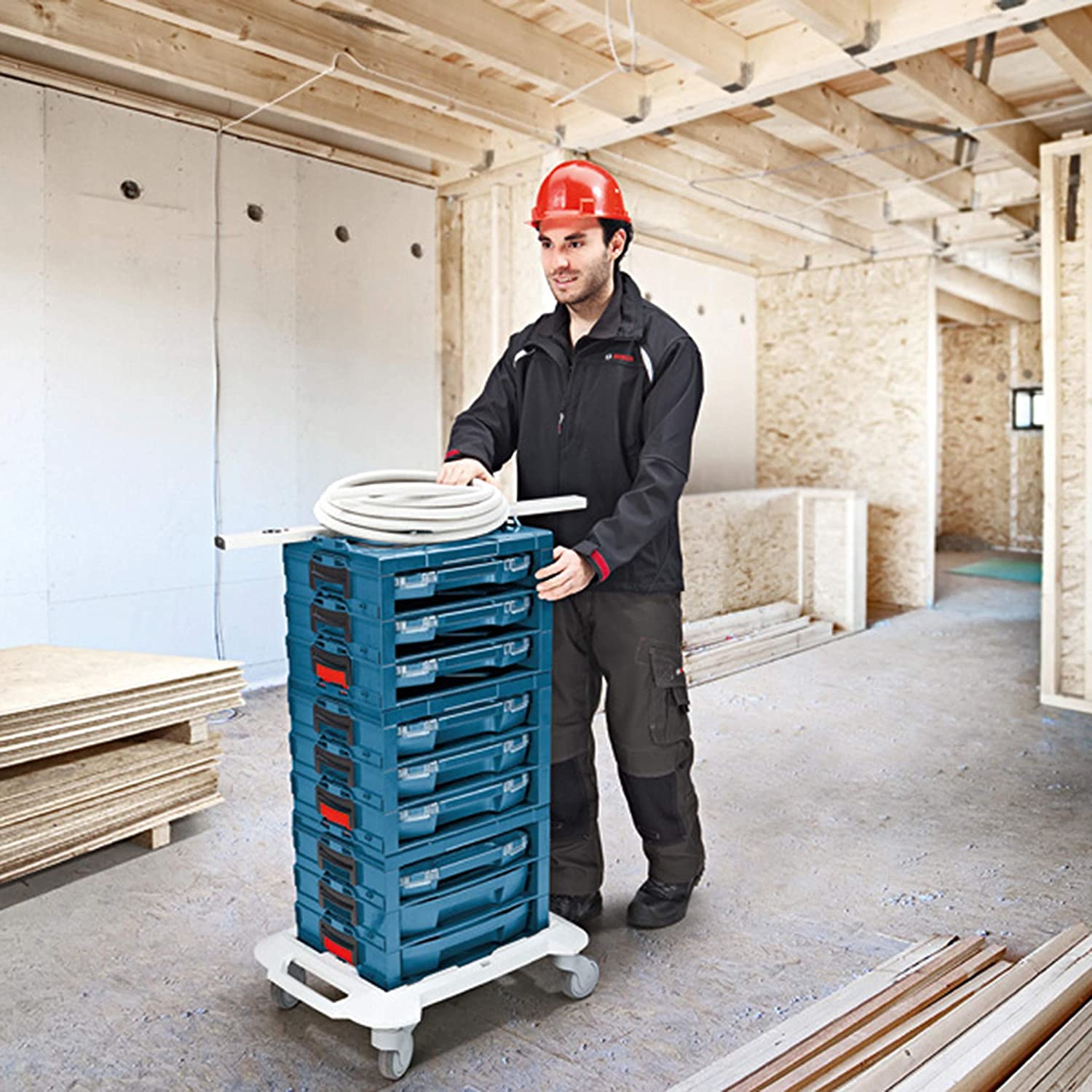 Mann in rotem Helm schiebt blaue Werkzeugkisten auf einem Rollwagen in einer Baustelle.