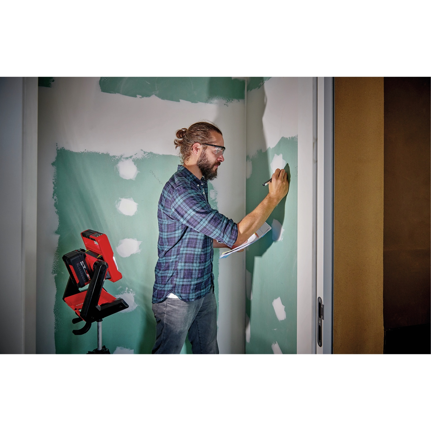 Mann mit Brille und kariertem Hemd markiert Wand. Rotes Licht auf Stativ.