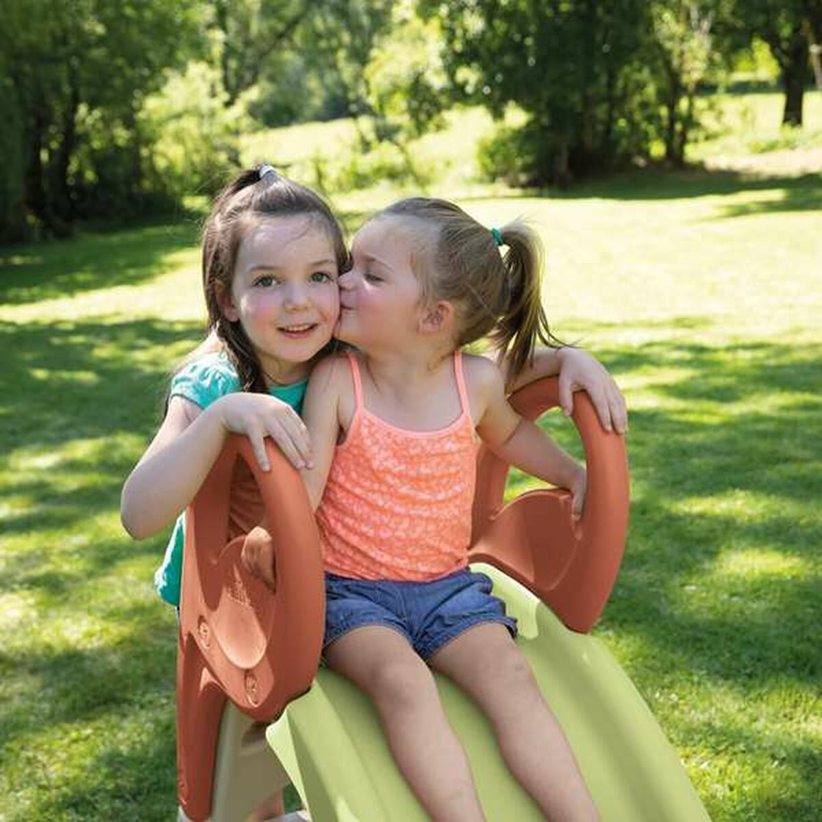 Due bambine su uno scivolo. Una bacia l'altra sulla guancia. Giornata di sole.