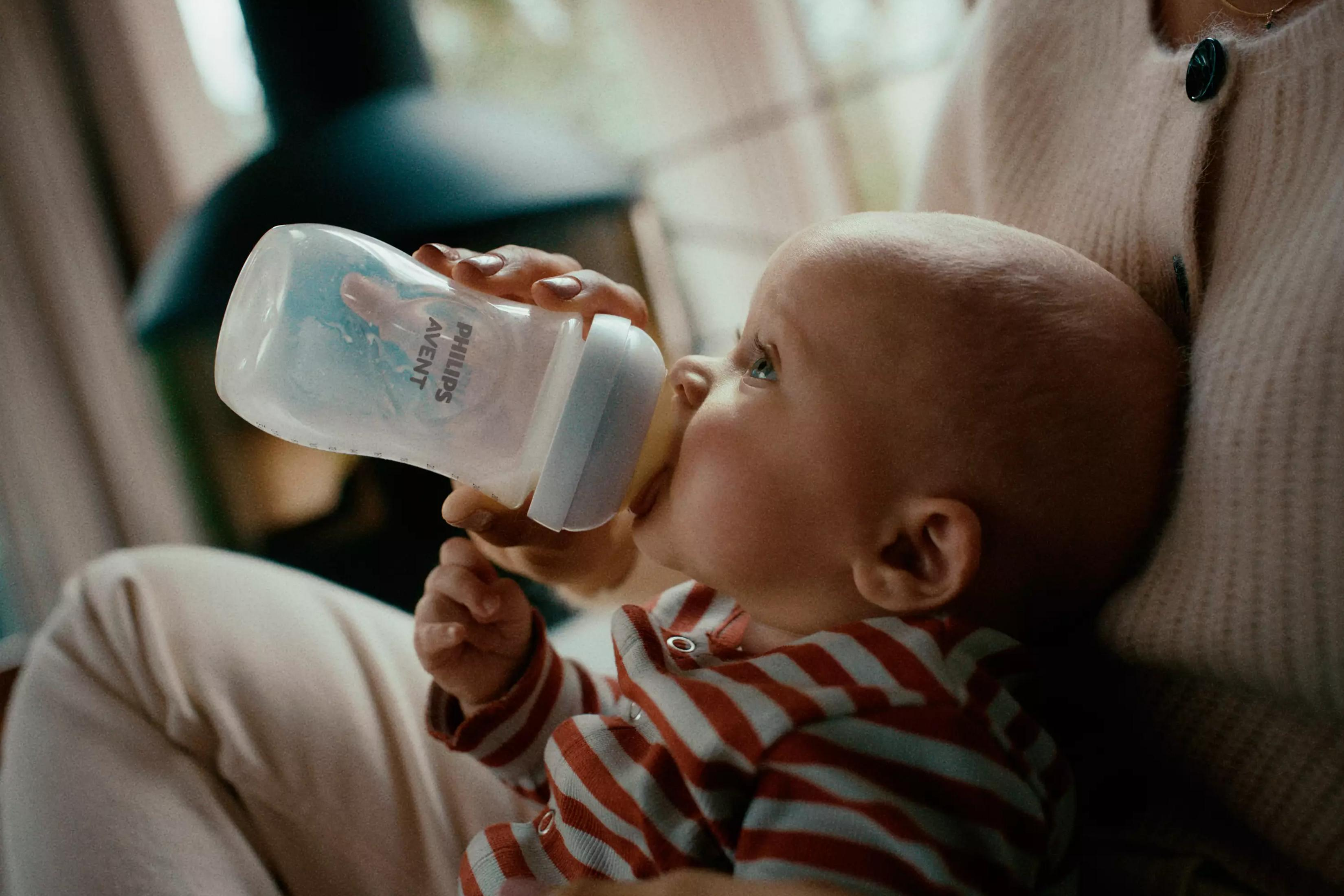 Ein Baby trinkt aus einer Philips Avent Flasche, getragen von einer Person in einem hellen Cardigan.