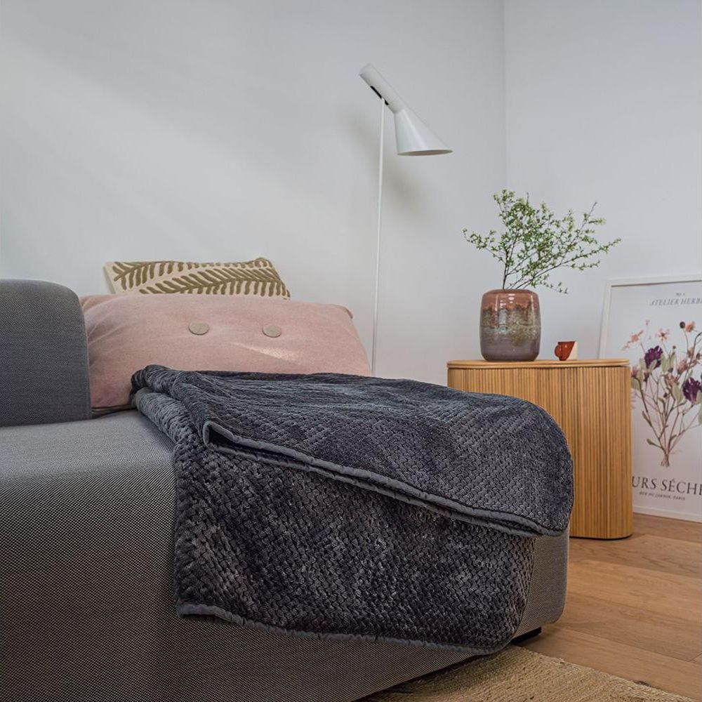 Ein dunkelgrauer Überwurf bedeckt ein graues Sofa mit rosa und grünen Kissen. Lampe und Vase sind in der Nähe.
