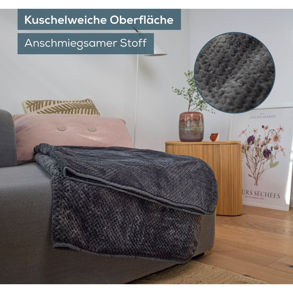 Ein dunkelgrauer Plüsch-Überwurf liegt auf einem Sofa, daneben Kissen und ein Beistelltisch.