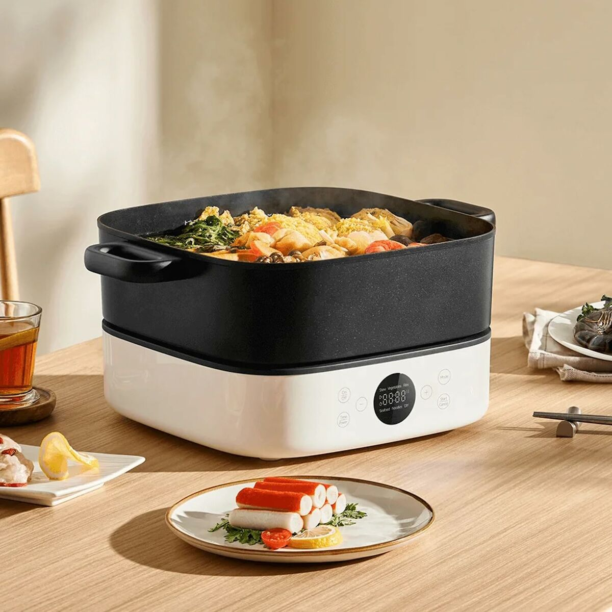 Una olla eléctrica de varios niveles cocina al vapor una comida de verduras y mariscos, con palitos de cangrejo en un plato.