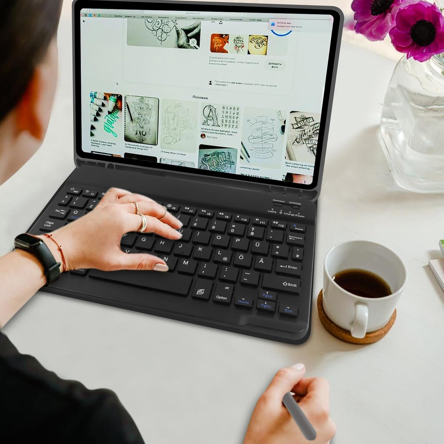 Eine Person tippt auf einer Tastatur, während sie einen Stift hält. Eine Tasse Kaffee steht daneben.
