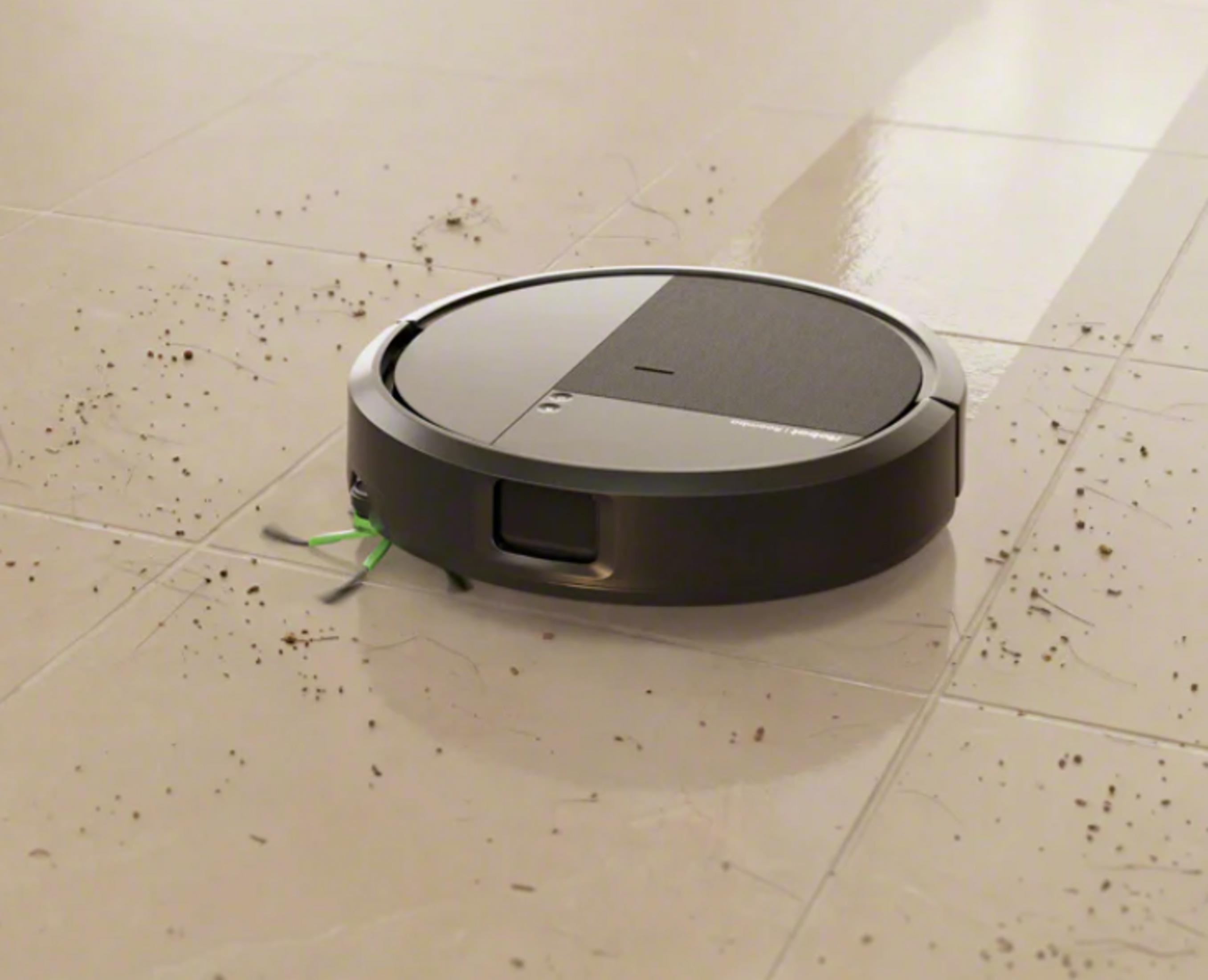 Un robot aspirador redondo con cepillos verdes sobre un suelo de baldosas sucio.