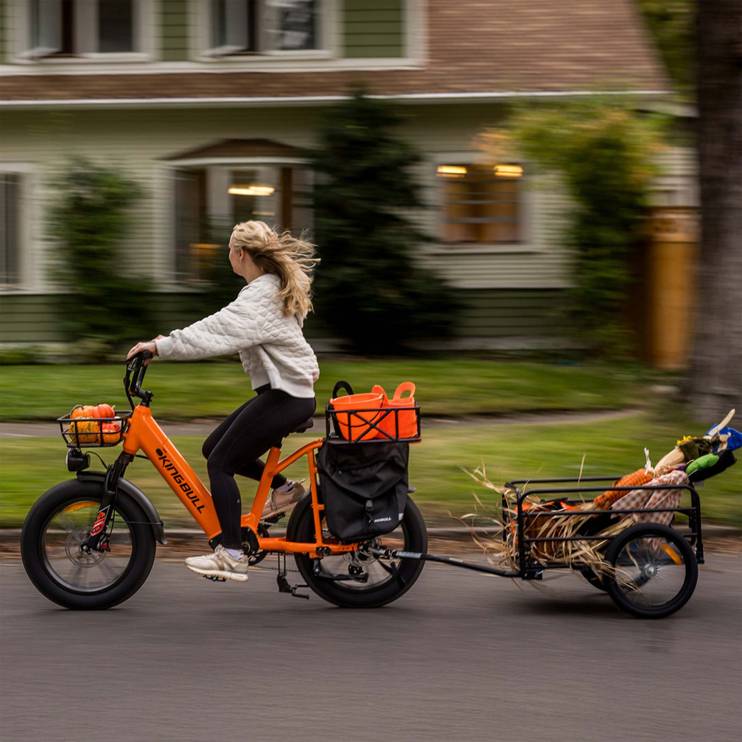 Eine Frau fährt ein orangefarbenes Fahrrad mit Anhänger, beladen mit Kürbissen und einer Vogelscheuche.