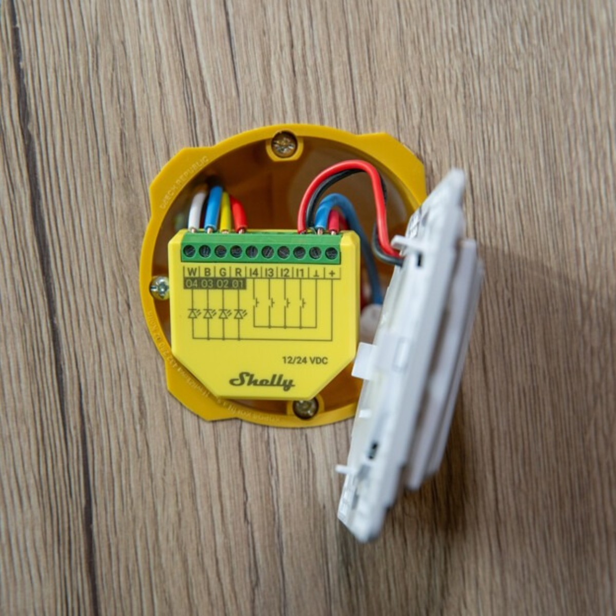 Żółty moduł Shelly z przewodami w puszce ściennej obok białego przełącznika.