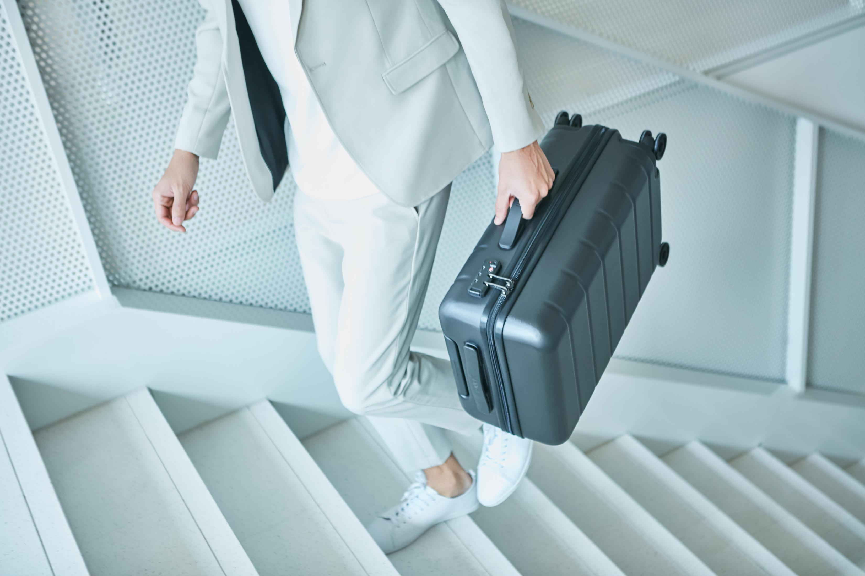 Una persona con ropa clara y traje blanco sube una maleta negra por las escaleras.