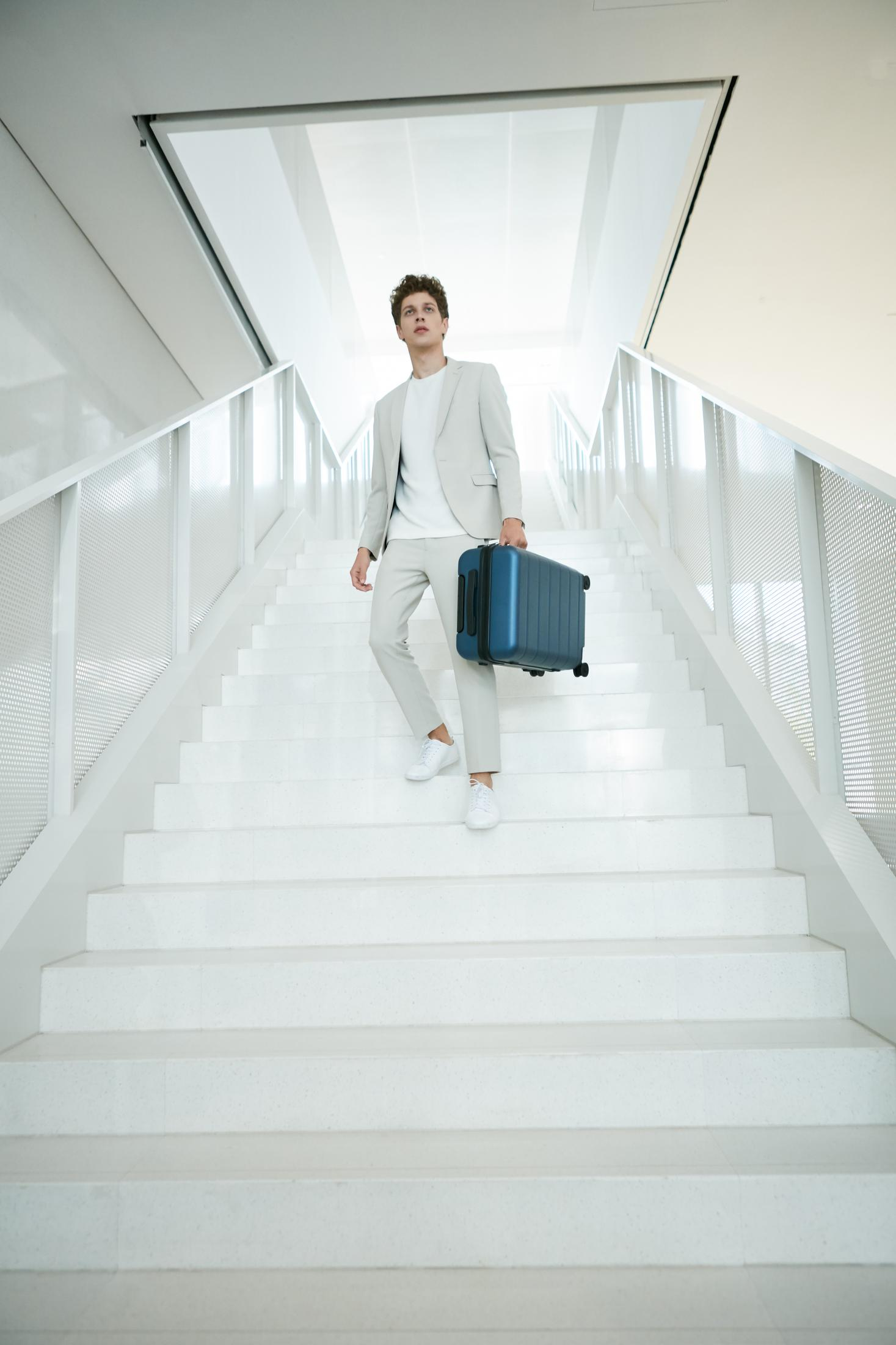 Un hombre con ropa beige lleva una maleta azul por una escalera blanca.