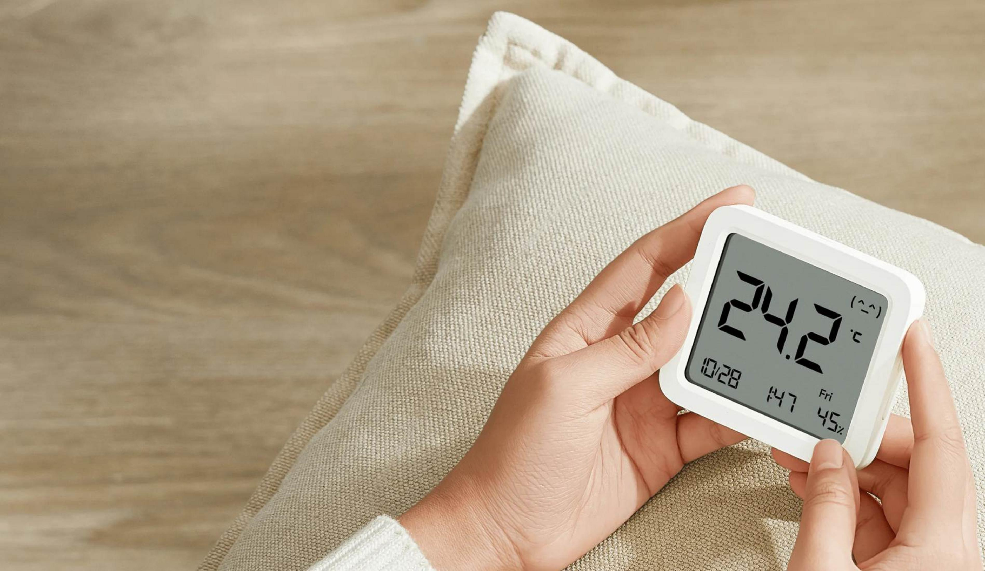 Una mano sostiene un termómetro blanco con temperatura, hora y humedad.