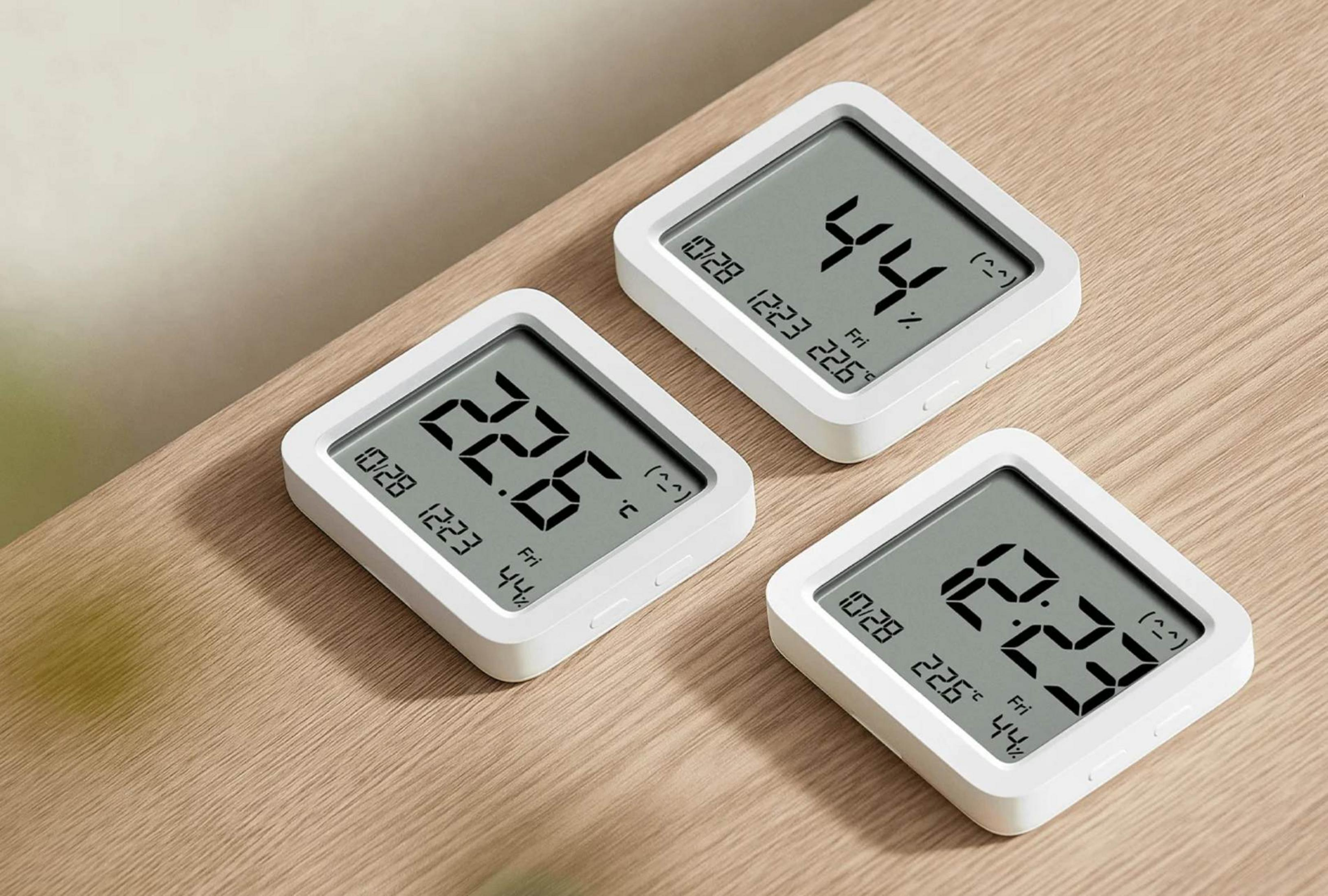 Tres termómetros/higrómetros digitales blancos sobre mesa de madera. Muestran hora, fecha, temp y humedad.