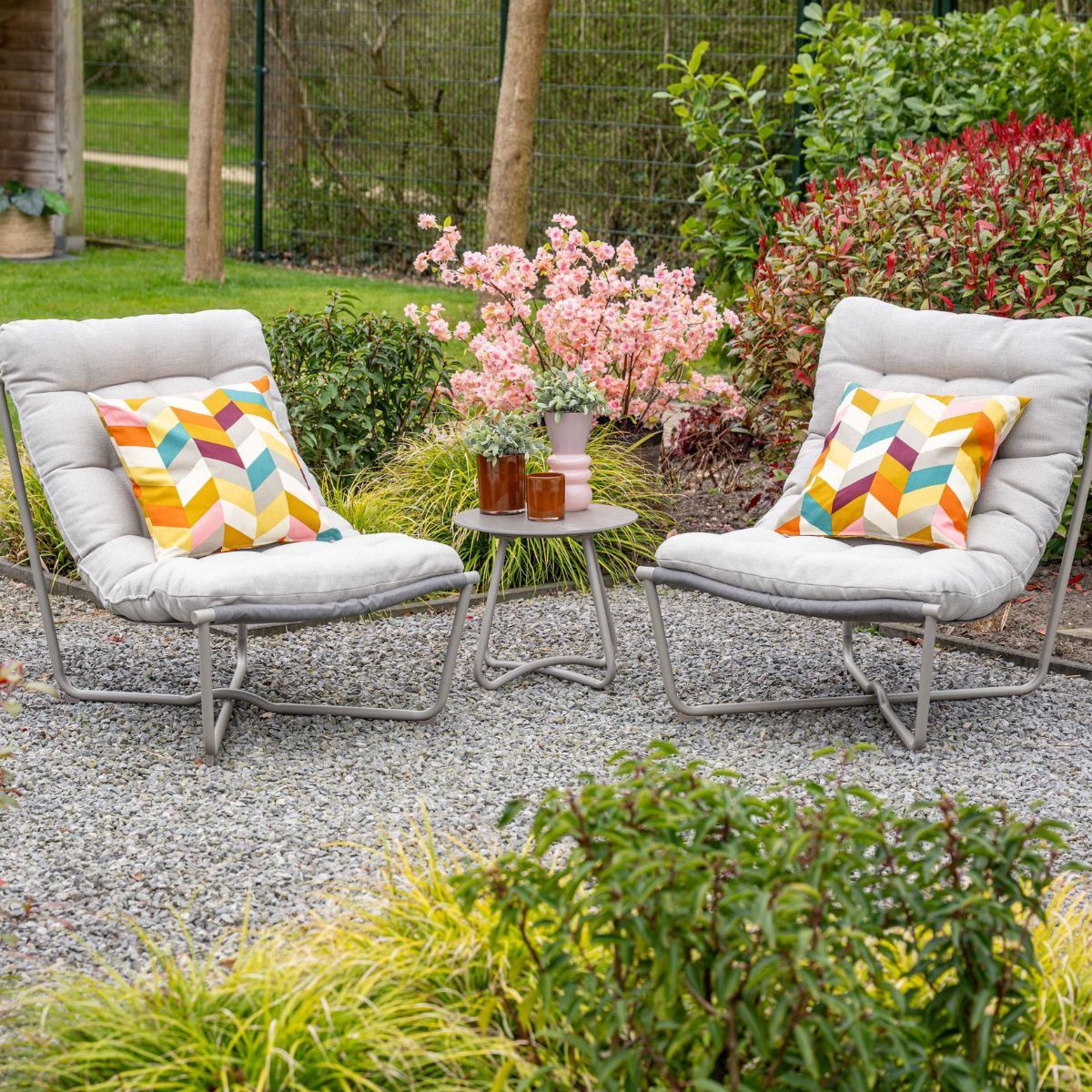 Two grey chairs with colorful pillows sit on gravel before blooming bushes.
