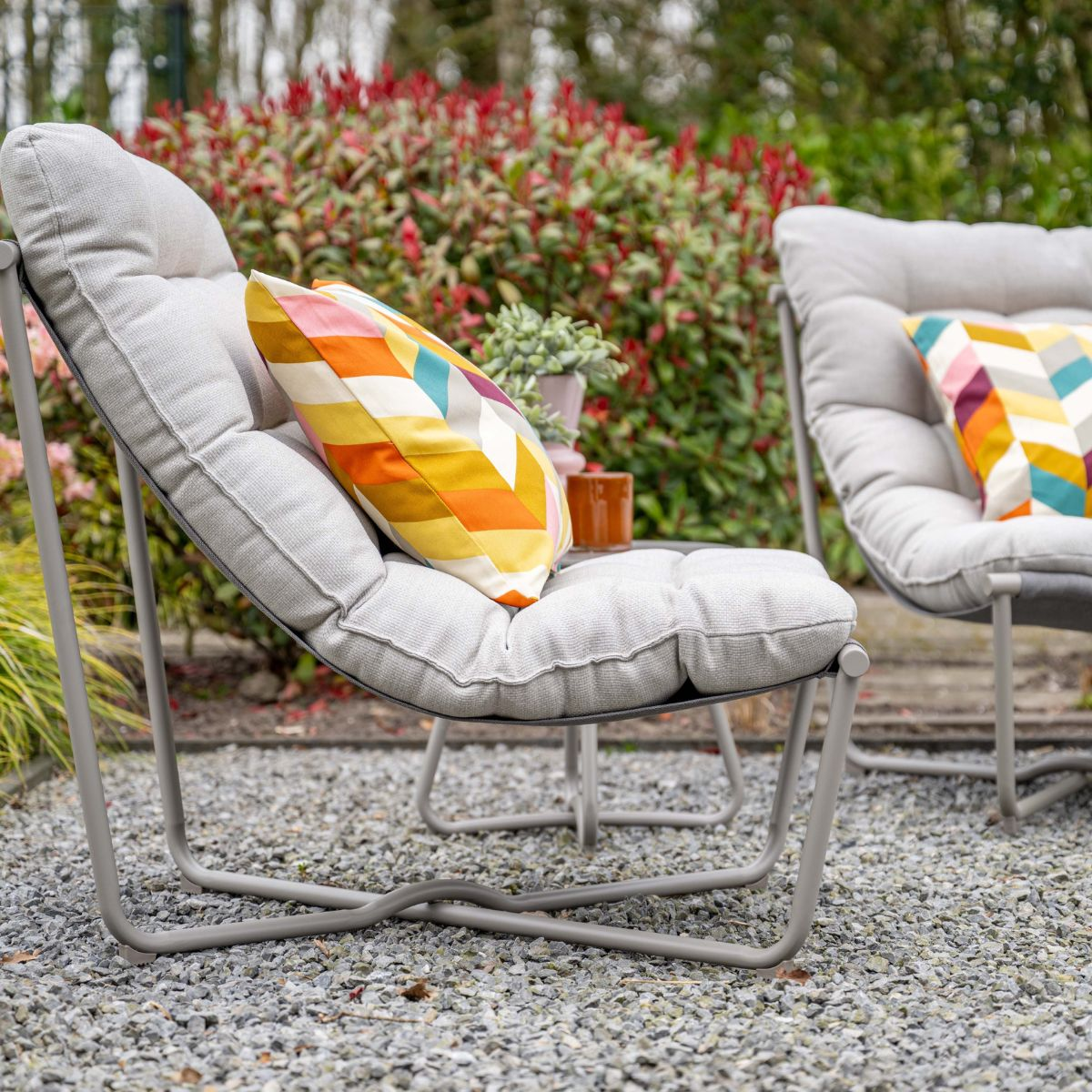 Two grey garden chairs with colorful cushions on gravel, red bushes in the background.
