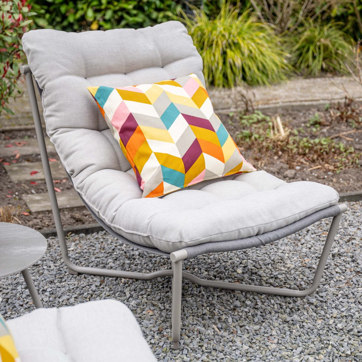 Gray lounge chair with colorful pillow on gravel ground in garden.