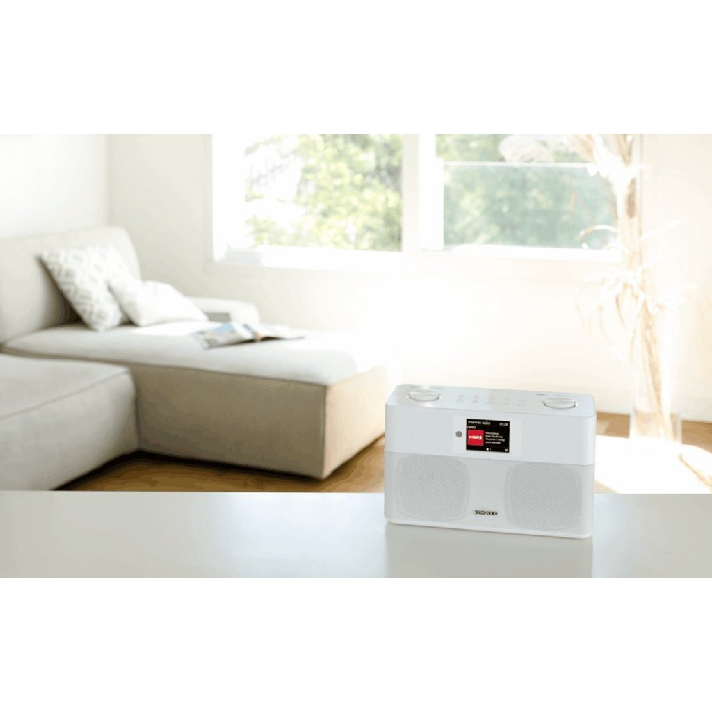 Ein weißes Radio mit Bildschirm steht auf einem Tisch vor einem Sofa und Fenstern.
