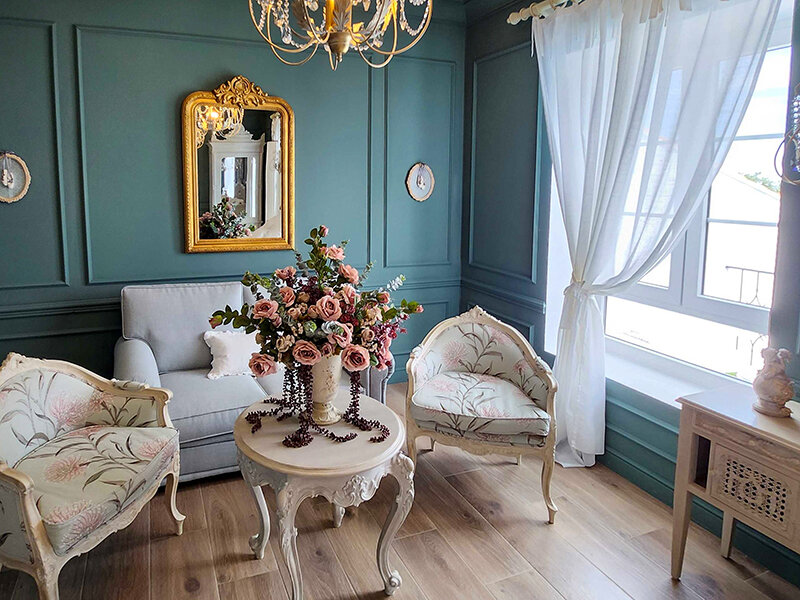 Una sala de estar vintage con sillones de felpa, un arreglo floral y un espejo dorado.