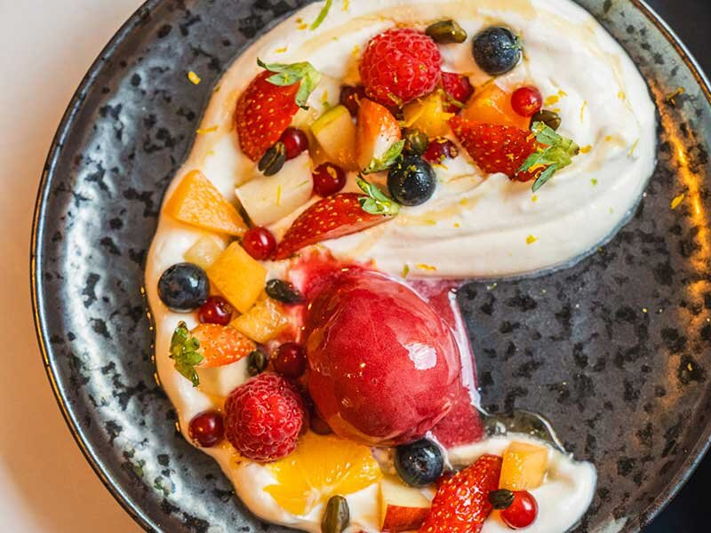 Postre cremoso con bayas, trozos de fruta y una bola de sorbete en plato oscuro.