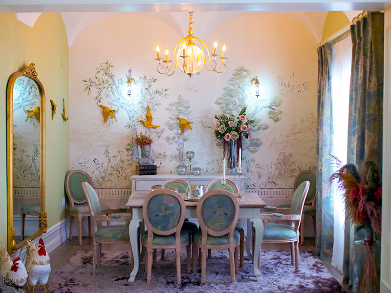 Un comedor con papel tapiz floral, mesa, sillas, espejo y figuras de pollos.