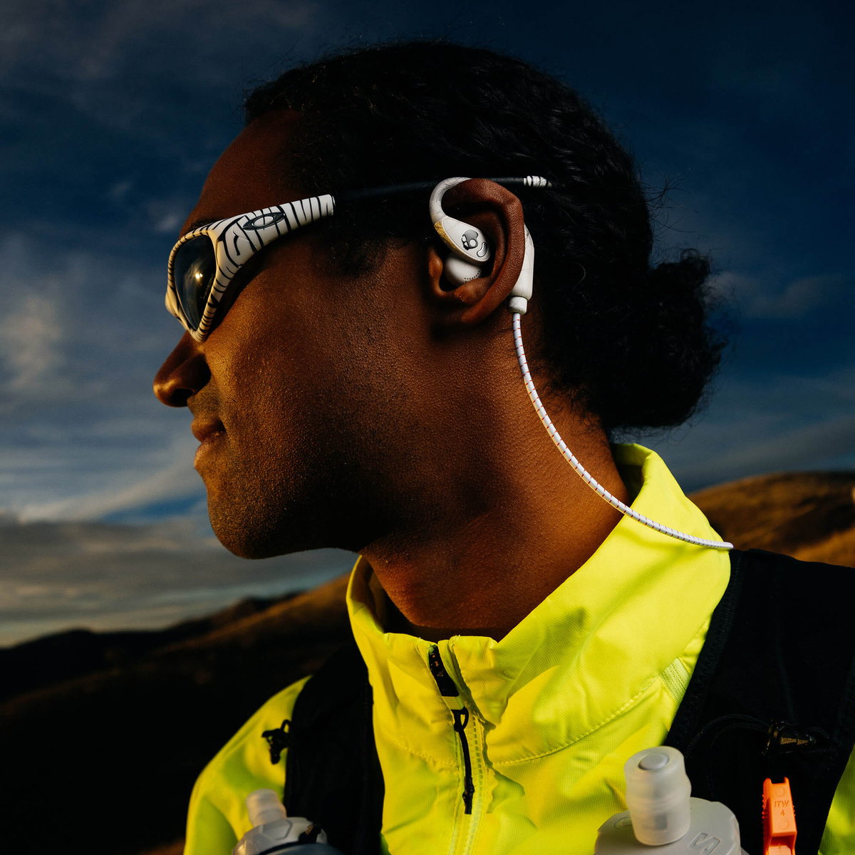 Mann mit Kopfhörern und Brille trägt eine gelbe Jacke, mit Berglandschaft im Hintergrund.
