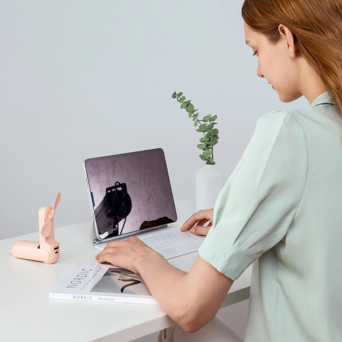 Una donna digita su un laptop. Una ventola rosa e una pianta sulla scrivania.