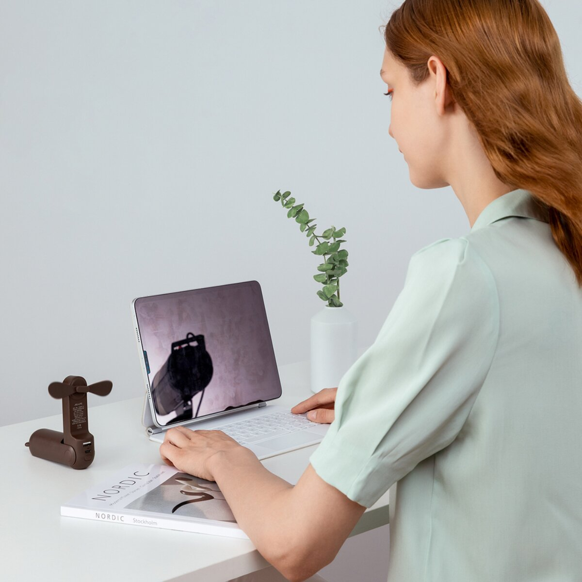 Eine Frau mit roten Haaren tippt auf einer Tastatur vor einem Tablet in einem hellen Raum.