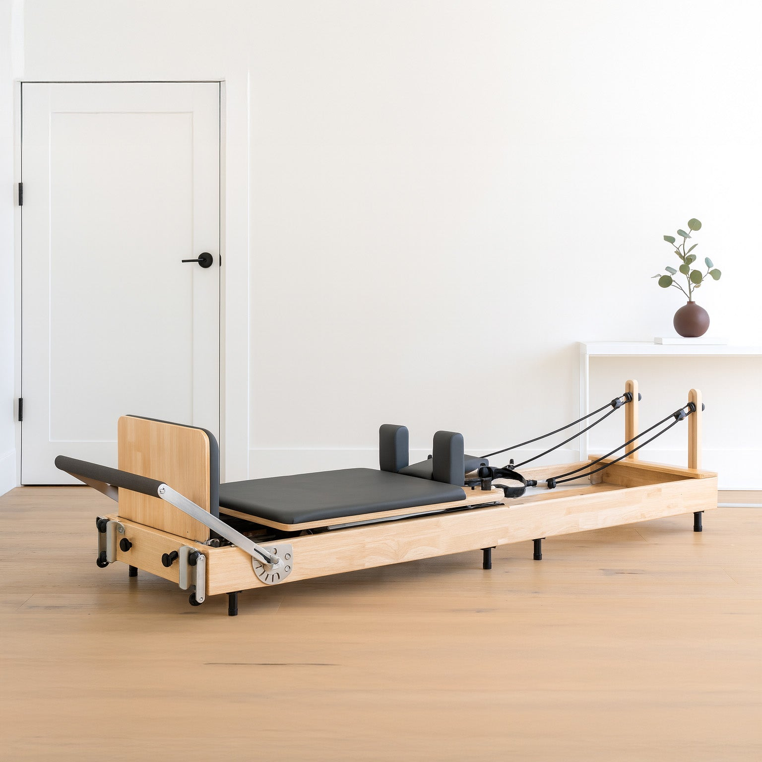 Un reformer de Pilates de madera con acolchado negro y cuerdas sobre suelo de madera.