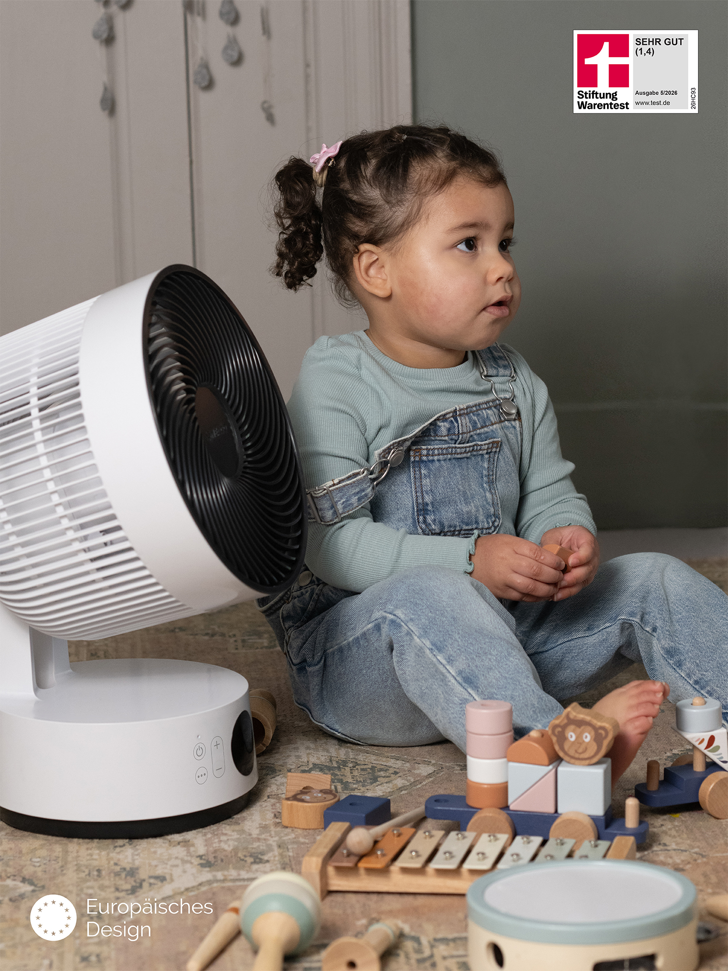 Ein kleines Kind sitzt auf dem Boden neben einem Ventilator und Spielzeug.