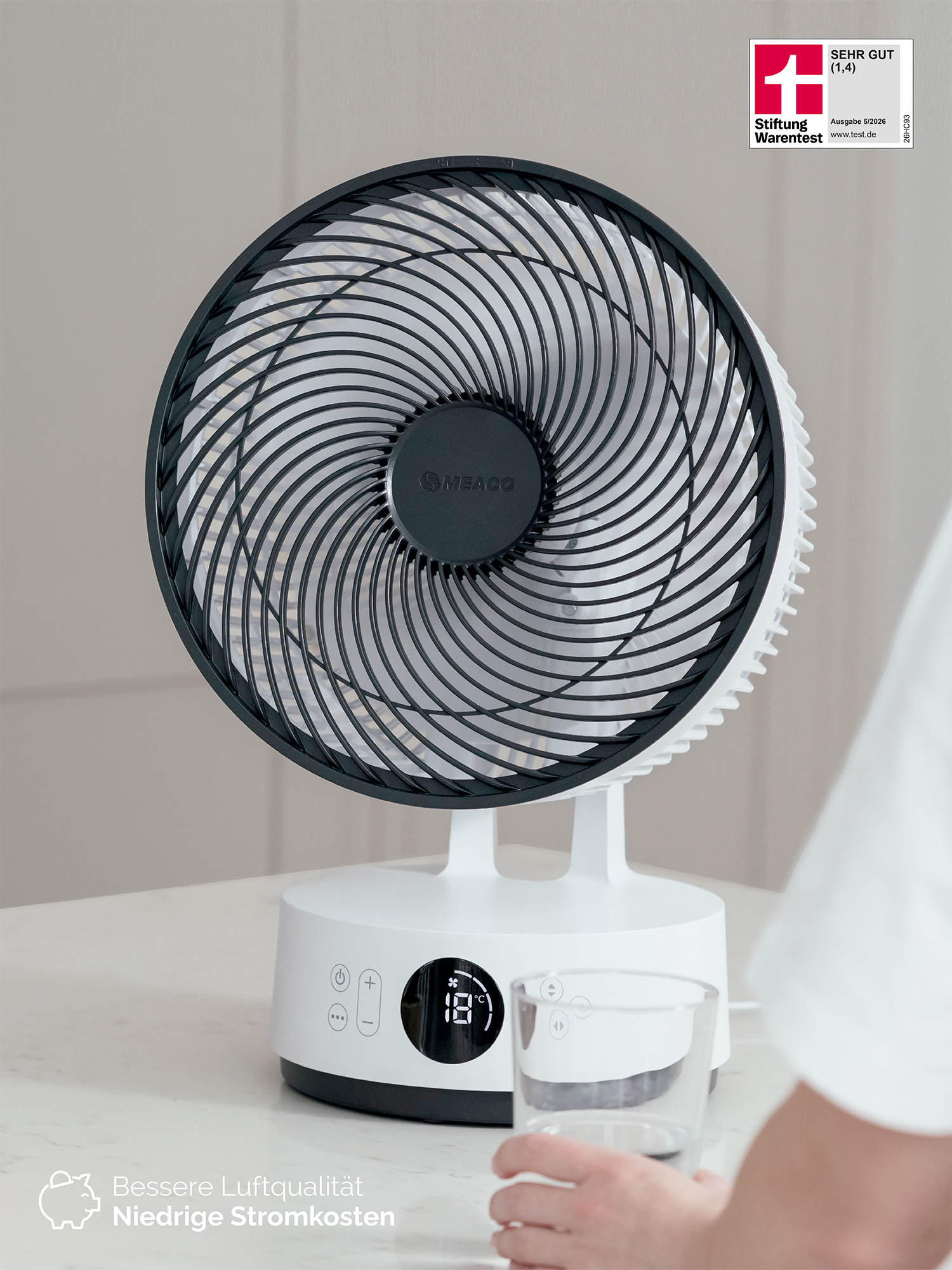 Ein weißer Ventilator mit schwarzem Gitter steht auf einem Tisch. Eine Hand hält ein Glas.