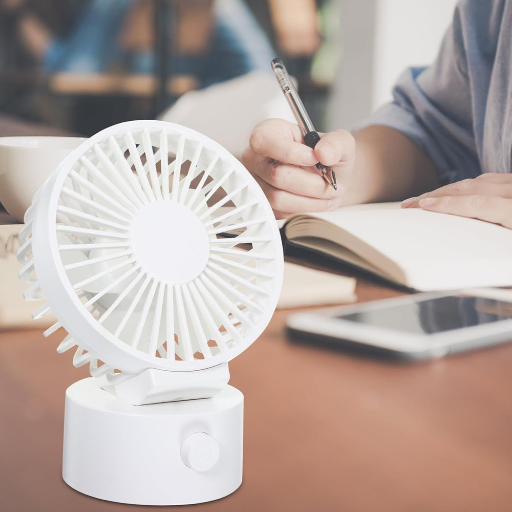 Ein kleiner weißer Ventilator steht auf einem braunen Tisch neben einem offenen Notizbuch und einem Stift.
